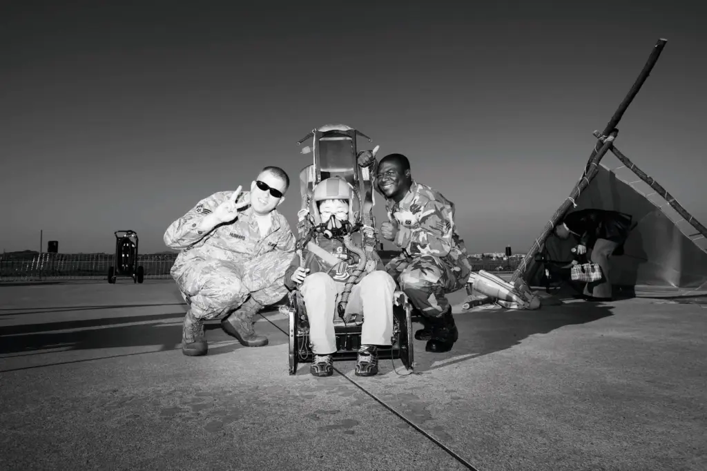 Ein Kind sitzt in Pilotenmontur in einem ausgestellten, ausgebauten Pilotensitz. Rechts und links davon posieren zwei Soldaten der US-Army und lächeln in die Kamera.