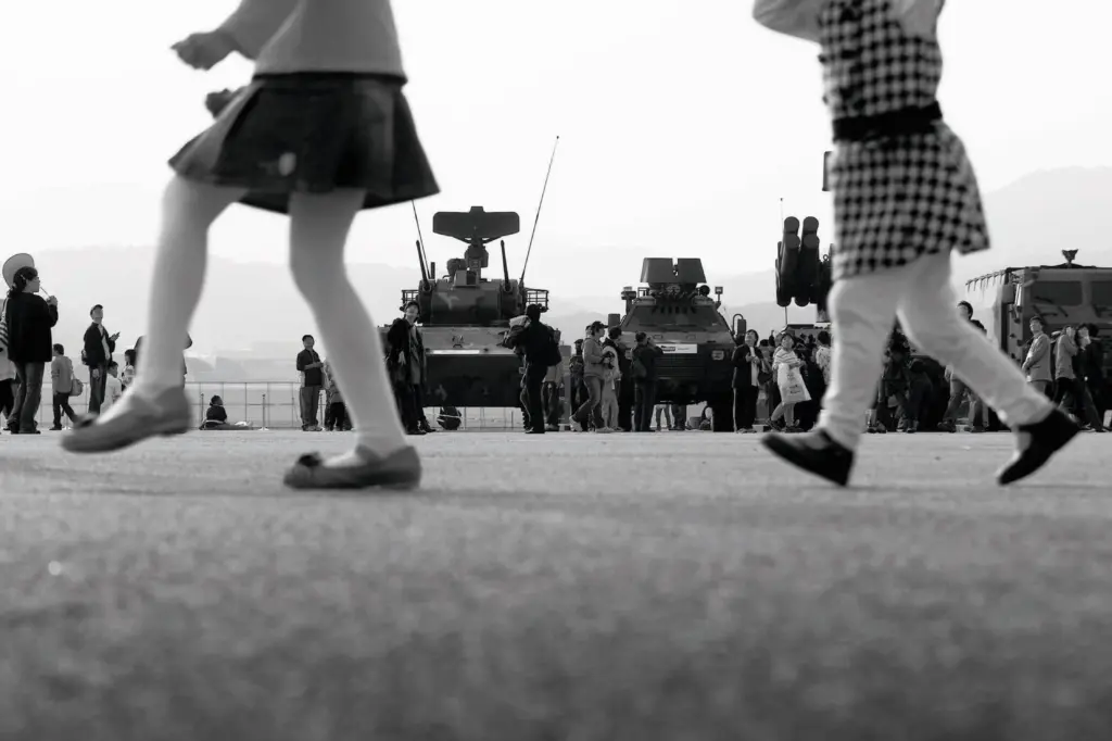 Zwei Kinder, von denen nur die Beine zu sehen sind, laufen an ausgestellten Militärfahrzeugen vorbei, die von zahlreichen Menschen begutachtet werden.