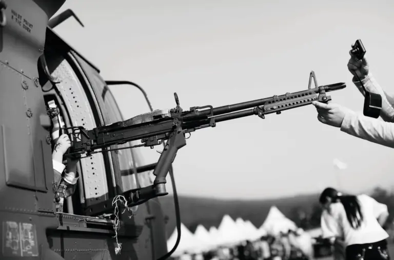 Ein Maschinengewehr ragt an der Seite eines Hubschraubers heraus. Am Abzug ist eine Kinderhand mit einer Kindertätowierung zu sehen, am Ende des Laufes schießt jemand ein Foto entlang des Laufes. Credit: Noh Suntag
