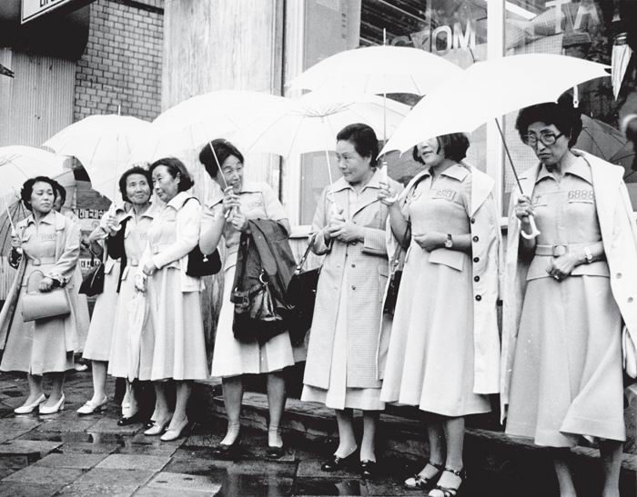 Schwarz-weiß Foto einer Gruppe von Frauen von Verhafteten des Myeongdong-Vorfalls in Trenchcoats und mit Regenschirm, die in einer Reihe stehend demonstrieren
